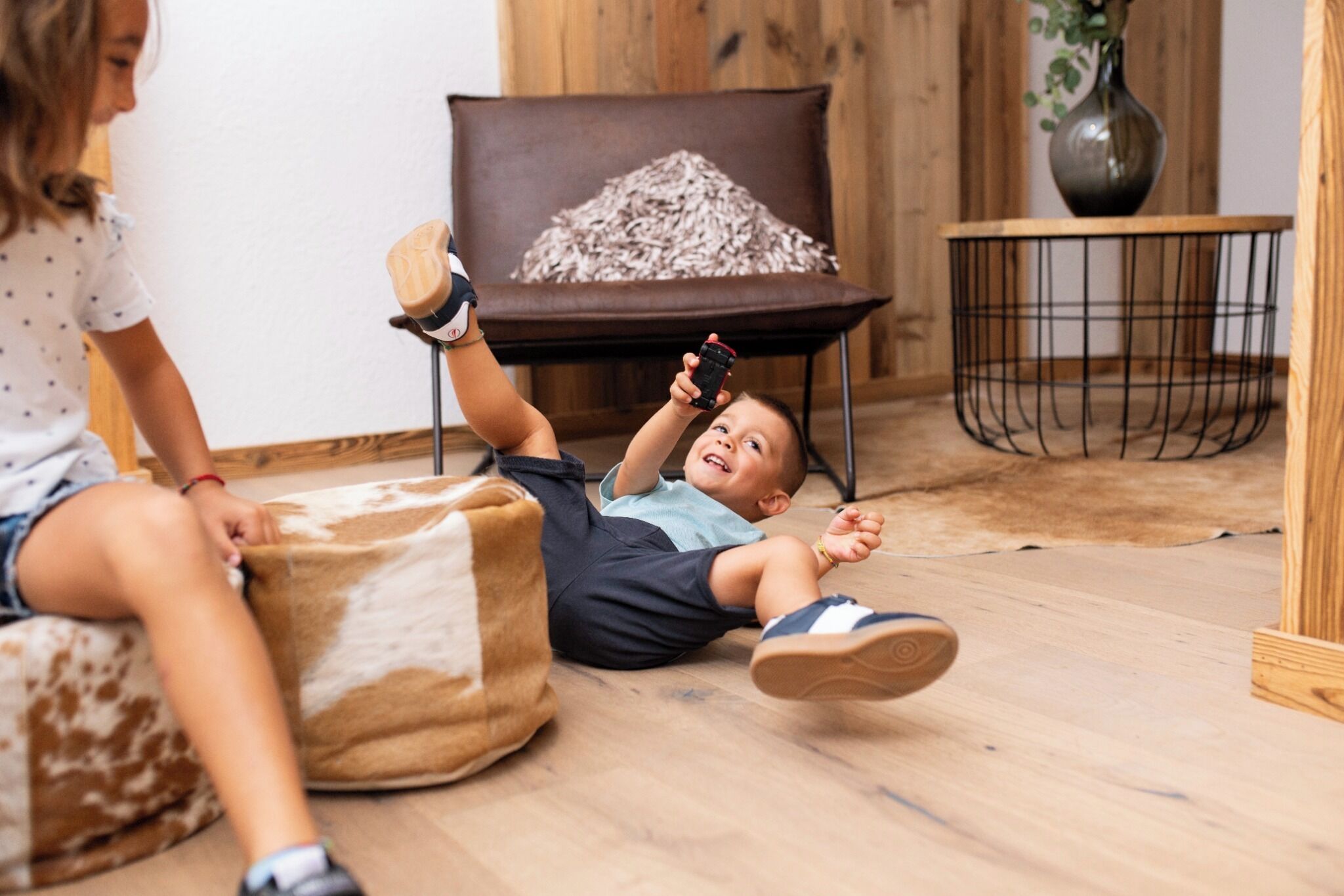 A child lying on the floor with a smartphone, a brown chair with a white cushion, and a small table with a vase.