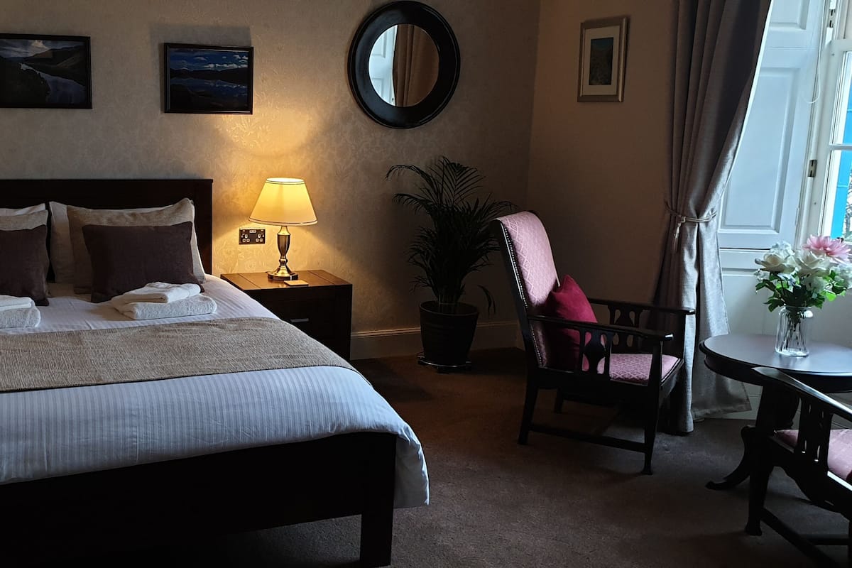 A neatly made bed with pillows and a blanket, two bedside lamps, a potted plant, a chair, and a table with a vase of flowers.