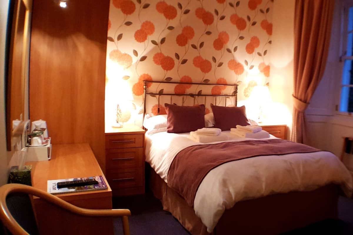 A small, cozy bedroom with a bed, bedside table, and a chair. The room has a floral wallpaper and a window with curtains.