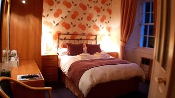 A small, cozy bedroom with a bed, bedside table, and a chair. The room has a floral wallpaper and a window with curtains.