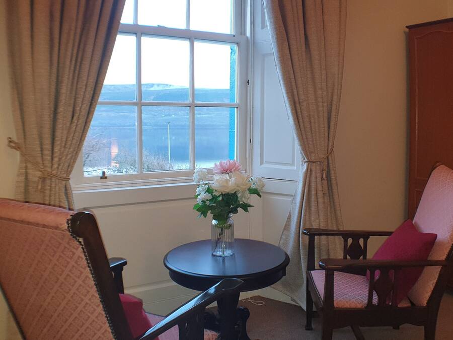 A room with a view of a landscape through a window, a round table with a vase of flowers, and two chairs.