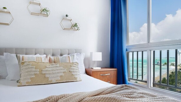 A bedroom with a bed, bedside table, lamp, and a view of the ocean through a balcony.