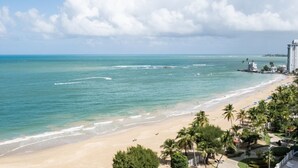 A beach with clear turquoise water, a sandy shore, and a tall hotel structure in the background.