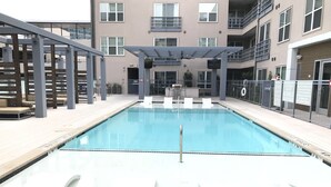 Una piscina con sillas de descanso y una pérgola, rodeada por un edificio de varios pisos con balcones.
