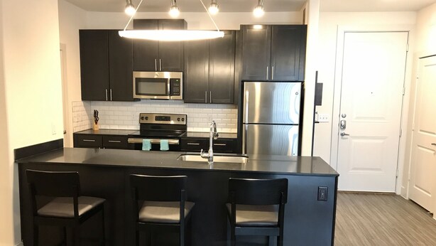 Una cocina moderna con armarios oscuros, electrodomésticos de acero inoxidable, una isla central con taburetes y luminarias colgantes.