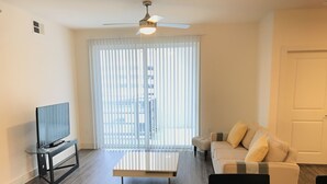 Una sala de estar con una televisión de pantalla plana sobre una mesa de vidrio, un sofá, una mesa de centro y un ventilador de techo.