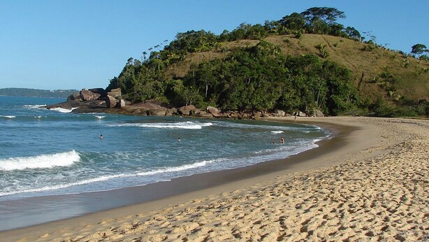 Une plage à l’eau d’un bleu limpide, avec un rivage sablonneux et une colline parsemée d’arbres.