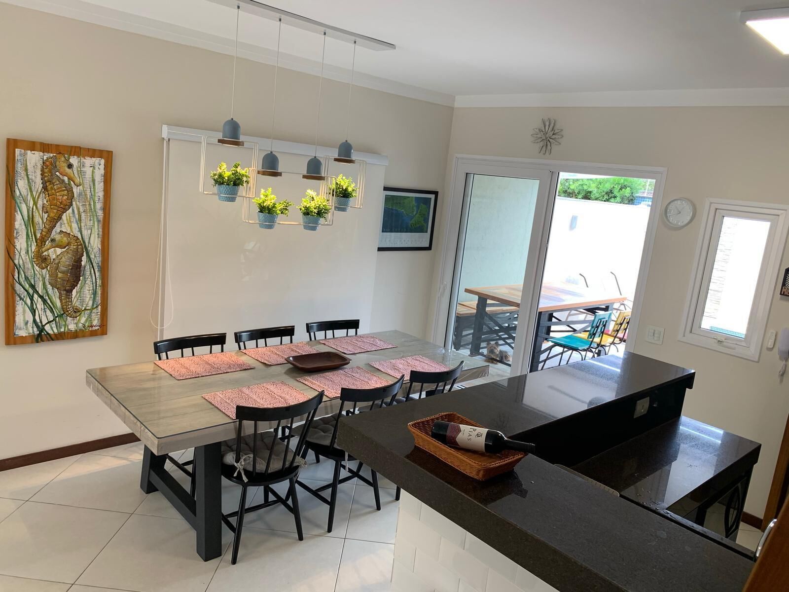 Uma cozinha moderna com uma ilha grande, cadeiras pretas e uma mesa de jantar posta para seis pessoas. Há plantas penduradas e um quadro na parede.