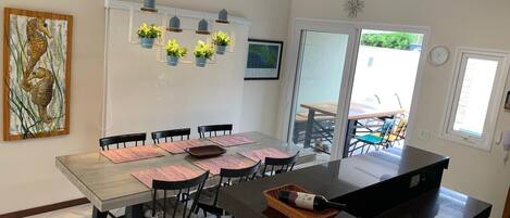 Uma cozinha moderna com uma ilha grande, cadeiras pretas e uma mesa de jantar posta para seis pessoas. Há plantas penduradas e um quadro na parede.