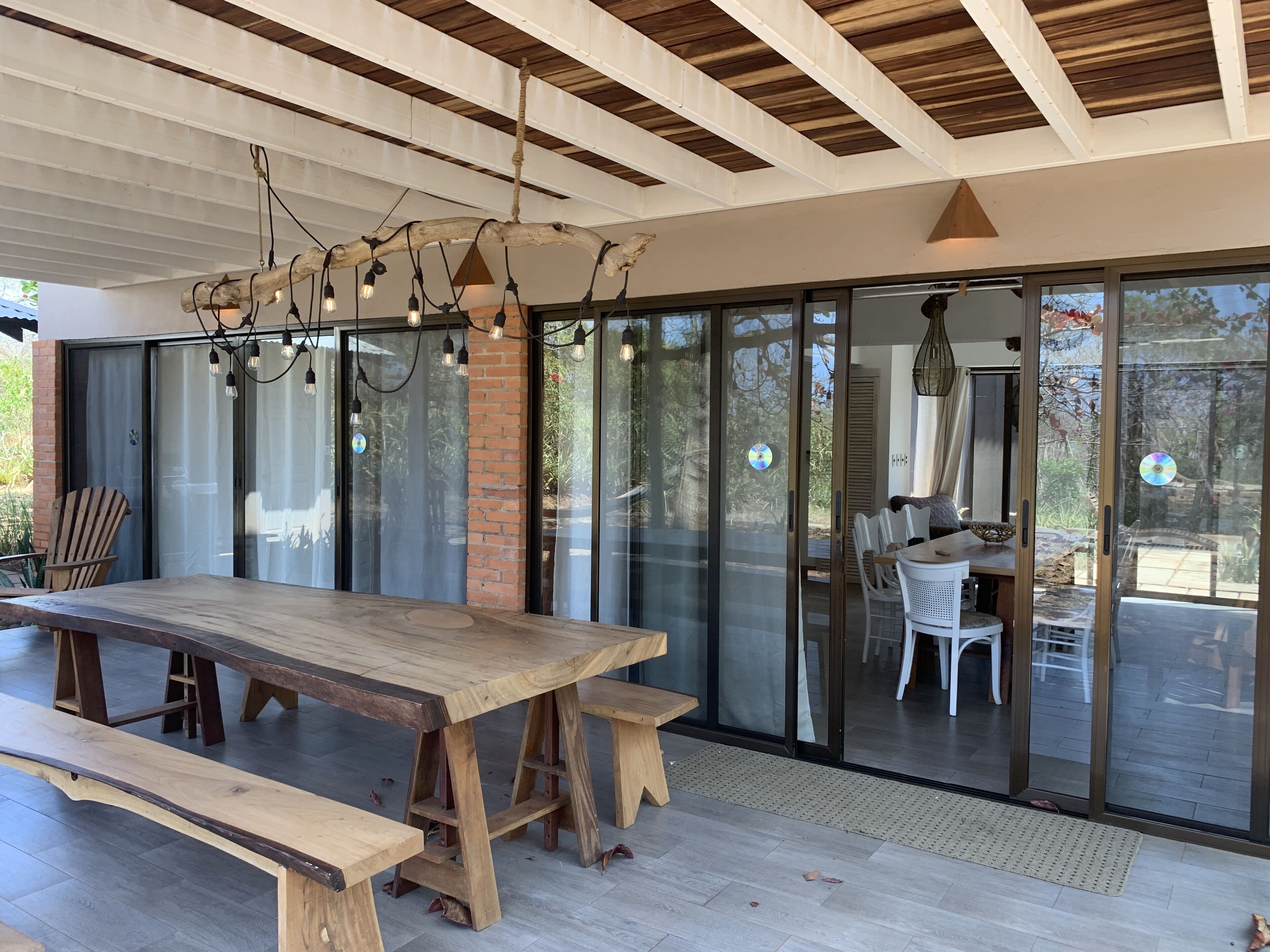 A spacious dining area with a large wooden table and benches, a hanging light fixture, and a view through large glass doors to an outdoor patio.