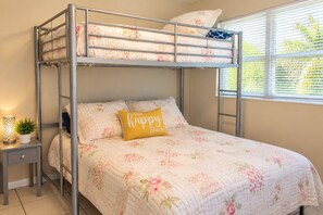 A bunk bed with a floral quilt, a bedside table with a lamp, and a window with blinds.