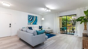 Een moderne woonkamer met een grijze bank, een witte fauteuil en een blauw vloerkleed. Er is een groot raam met uitzicht op groen.
