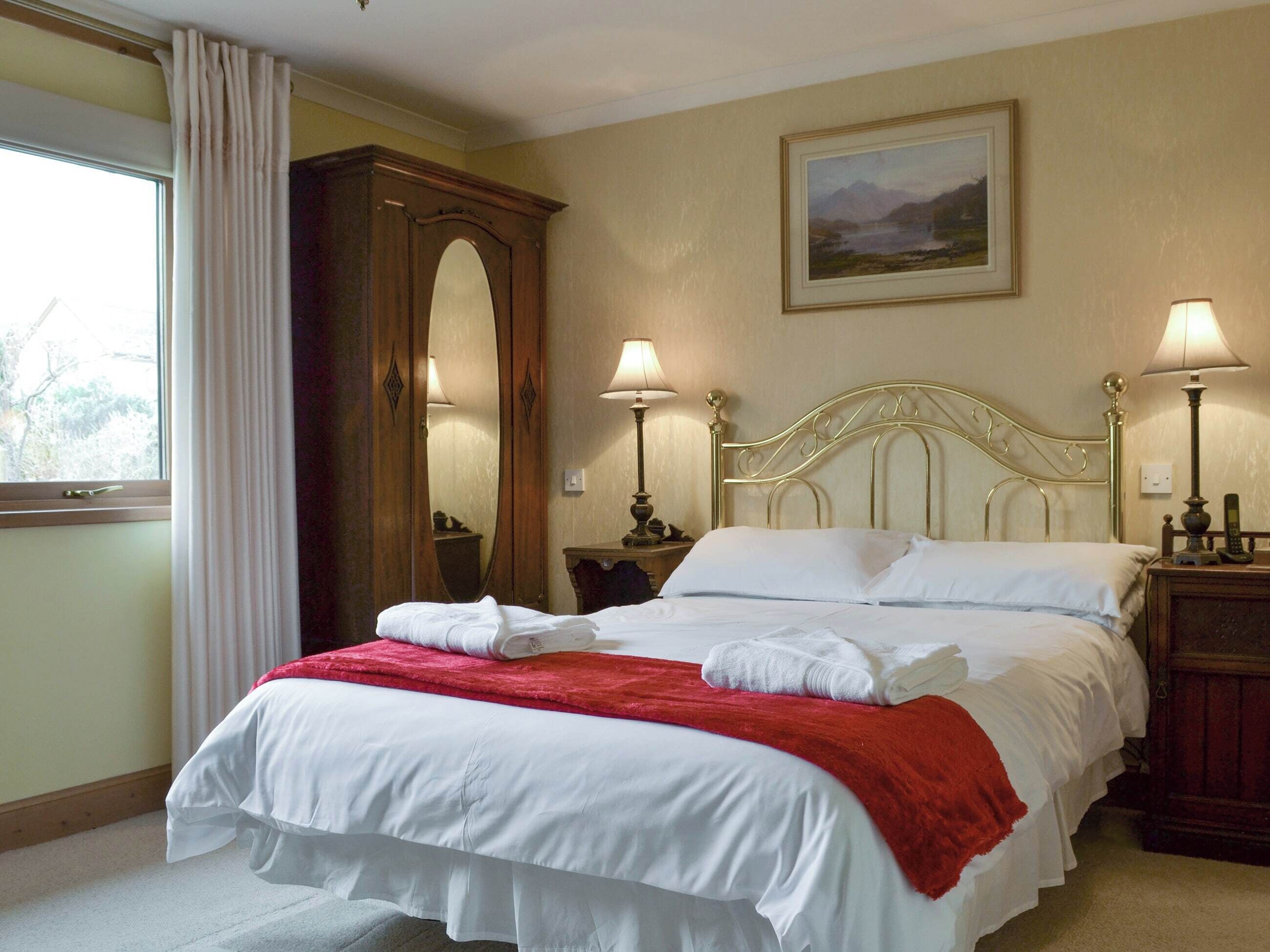 Un lit bien fait, avec du linge de lit blanc et une couverture rouge, deux lampes de chevet, une table de chevet en bois, un grand miroir et un tableau représentant un paysage, accroché au mur.