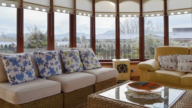 Ein gemütliches Zimmer mit Sofa und Sessel, große Fenster mit Blick auf Berge und Bäume sowie ein dekorativer Teller auf einem Glastisch.