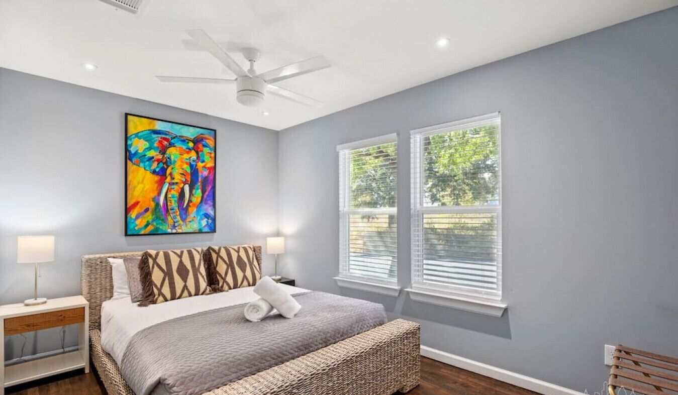 A bedroom with a bed, a ceiling fan, a painting, and two windows.