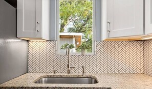 Enjoy a kitchen with a herringbone-patterned backsplash, granite countertop, and a stainless steel sink under a window with a view of the surrounding greenery.