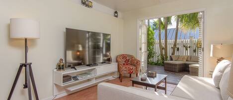 A living room with a TV, sofa, coffee table, and a chair. There is a balcony with a view of palm trees and a white fence.