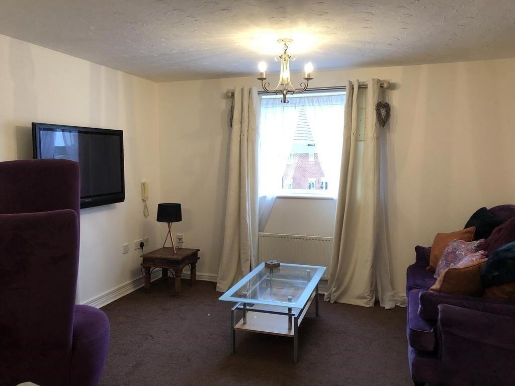 A living room with a glass-top coffee table, a purple armchair, a sofa, a flat-screen TV, and a window with curtains.
