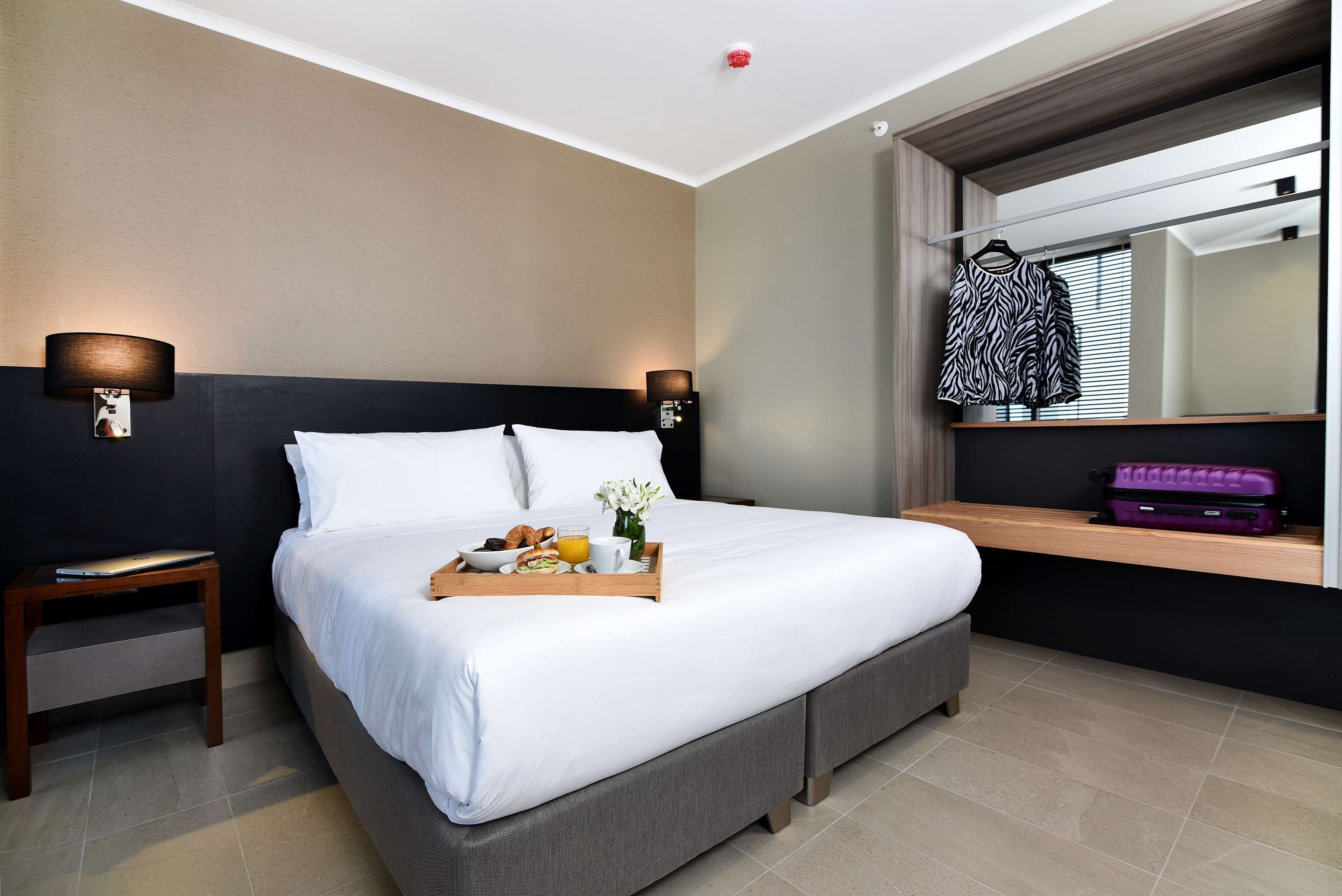 Een hotelkamer met een bed, nachtkastje, lamp en een plank met een koffer.