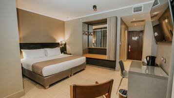 A hotel room with a bed, bedside table, lamp, mirror, wardrobe, and a small kitchenette.