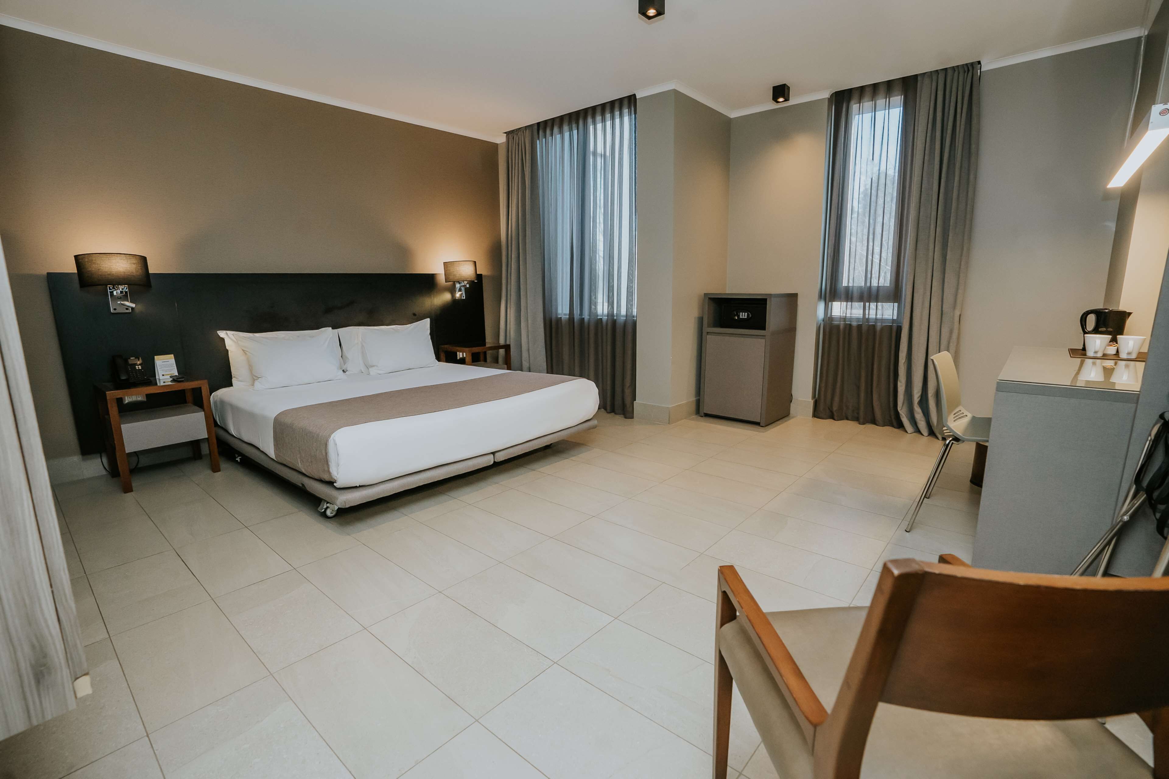 A hotel room with a large bed, bedside tables, a desk, a chair, and curtains.