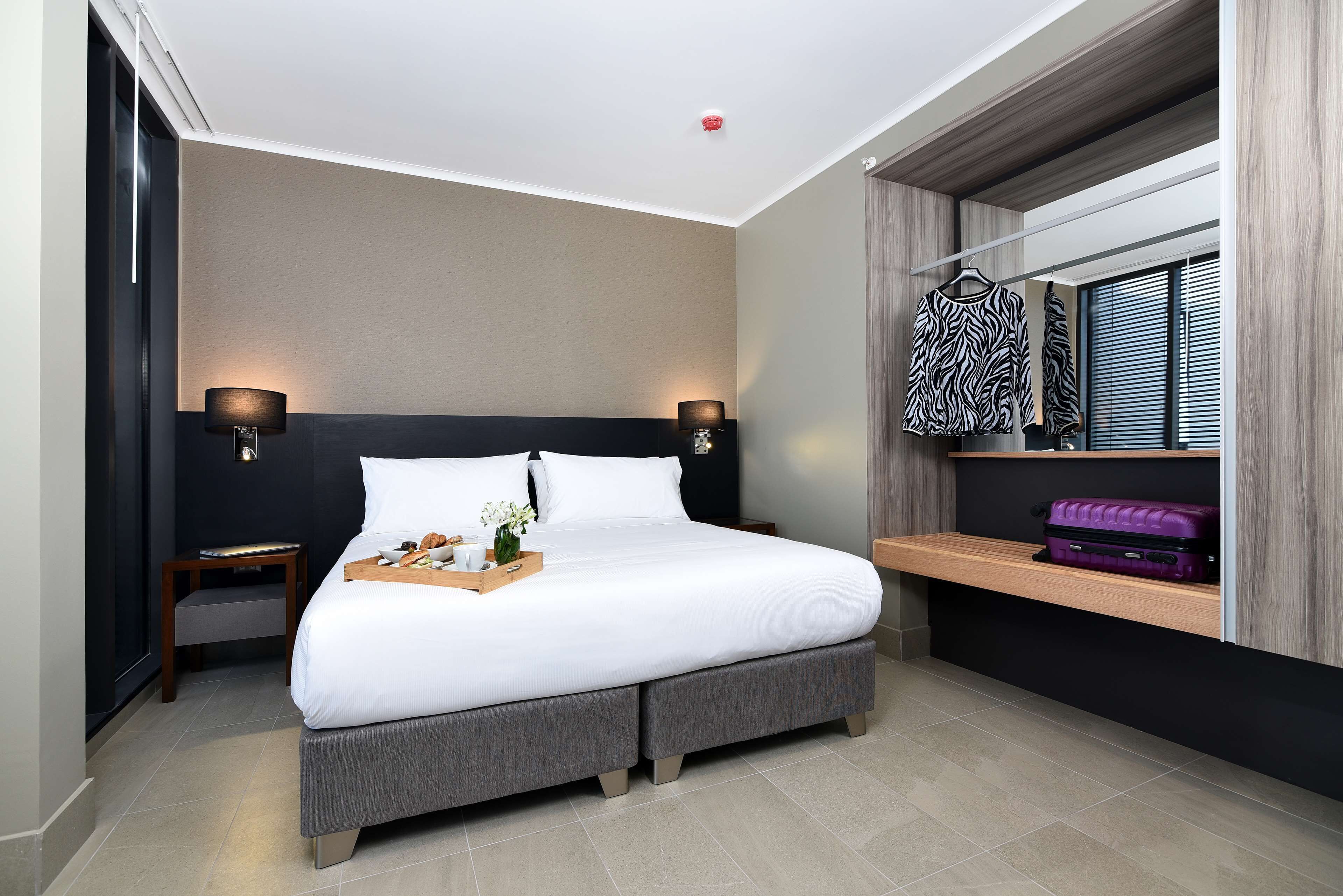 Ein modernes Hotelzimmer mit einem großen Bett, Nachttischlampen, einem kleinen Tisch mit Tablett und einem Fenster mit Jalousien.