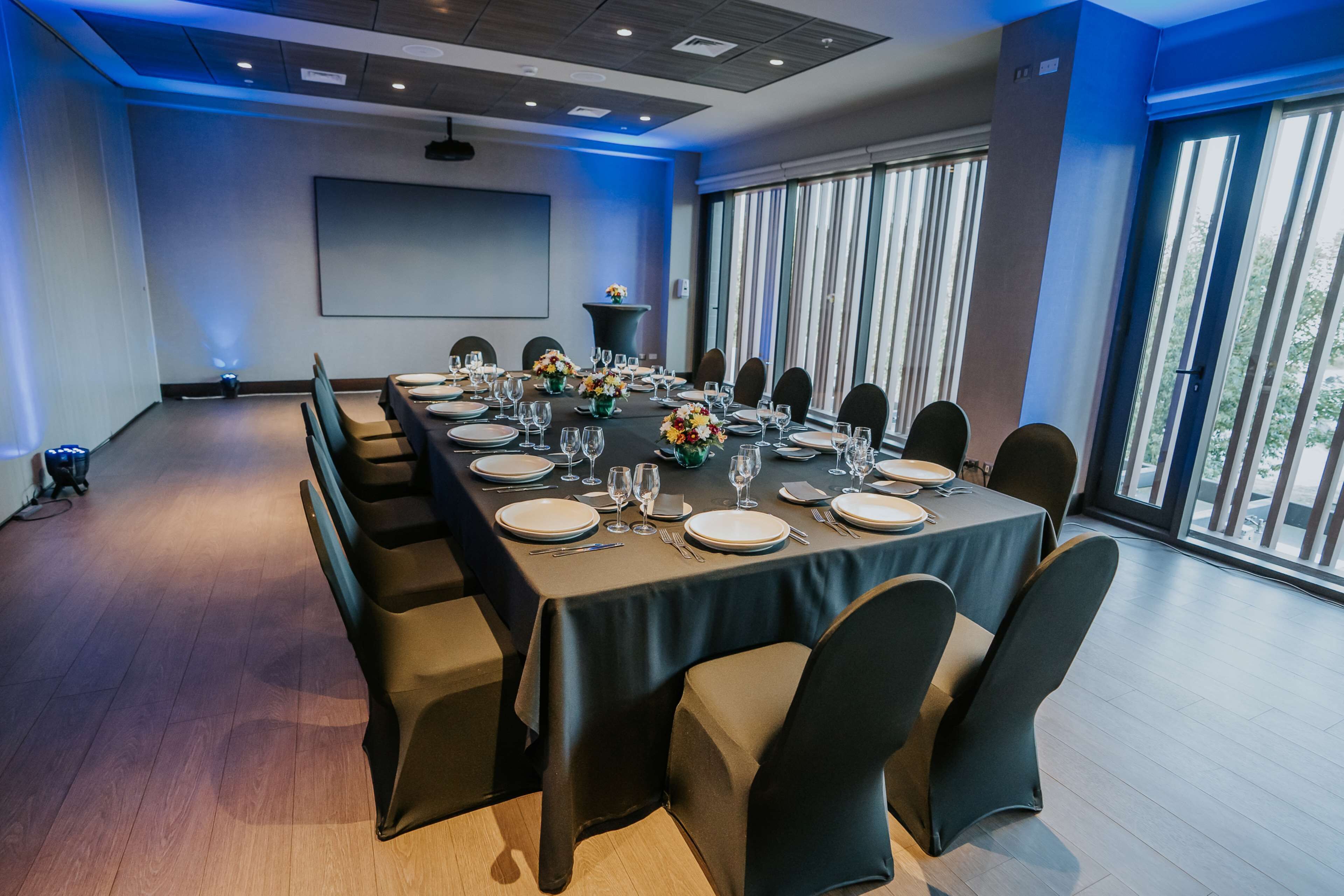 Un salón de conferencias con una larga mesa dispuesta para una reunión, ventanas amplias y una pantalla de proyector.