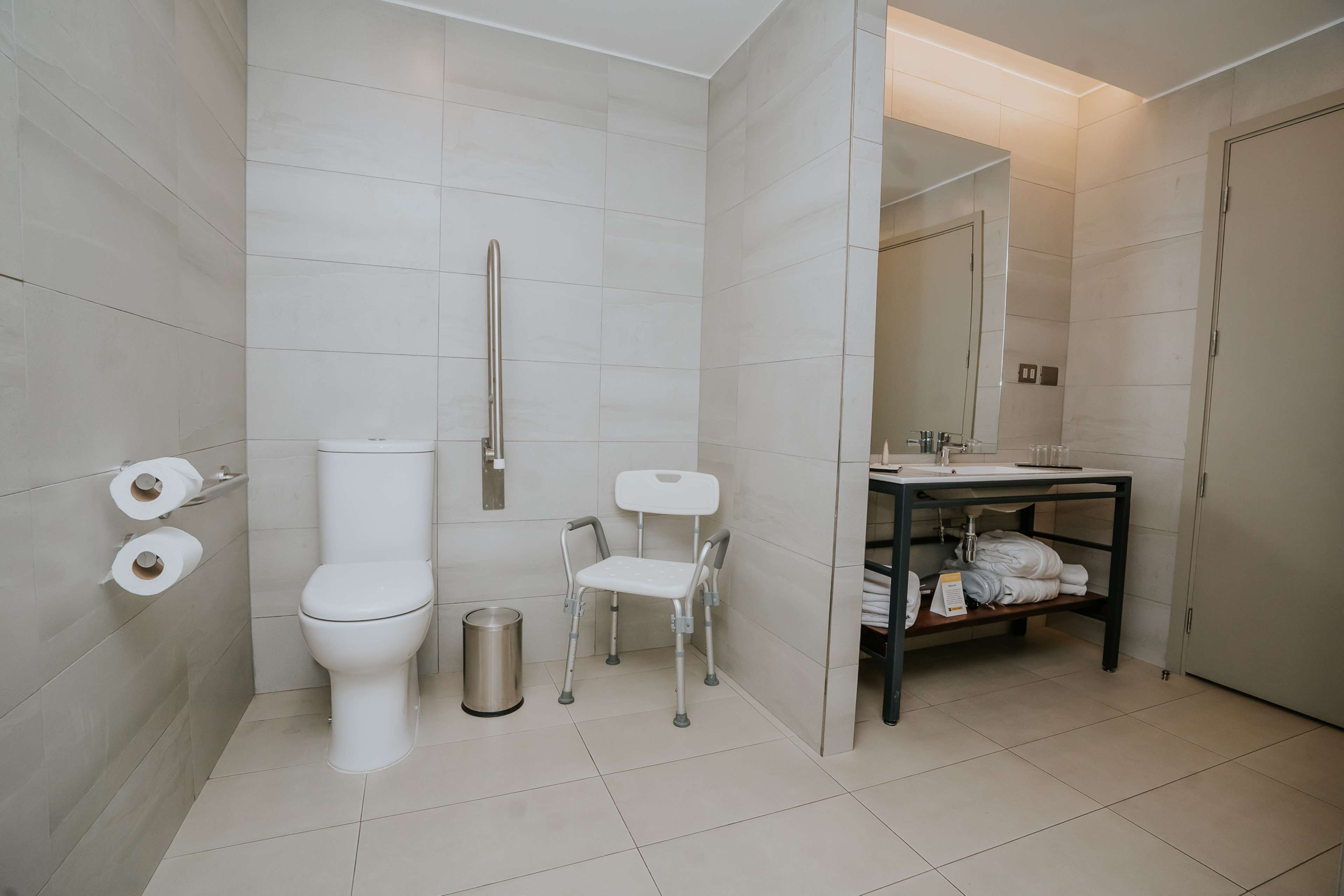 Un baño moderno con inodoro, bidé y una pequeña silla. Hay un lavamanos con un espejo encima y un estante con toallas.