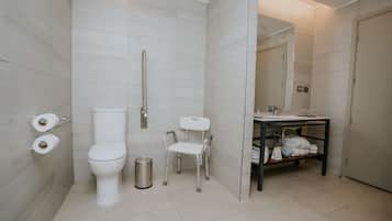 Un baño moderno con inodoro, bidé y una pequeña silla. Hay un lavamanos con un espejo encima y un estante con toallas.