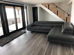 Een moderne woonkamer met een hoekbank, houten vloeren, een trap en een grote schuifdeur die leidt naar een buitenruimte.