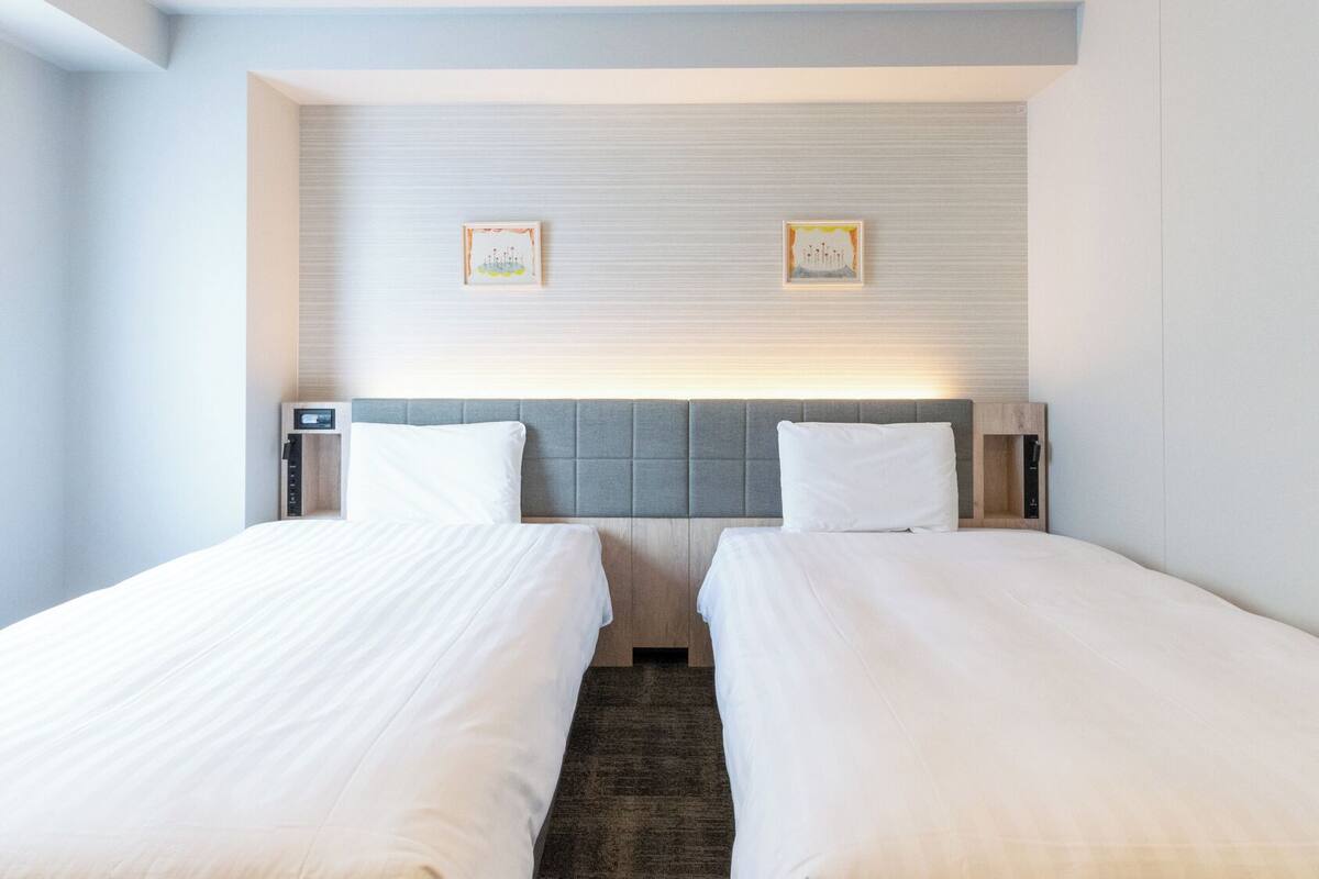 A hotel room with two beds, a headboard with a light panel, and two framed pictures on the wall.