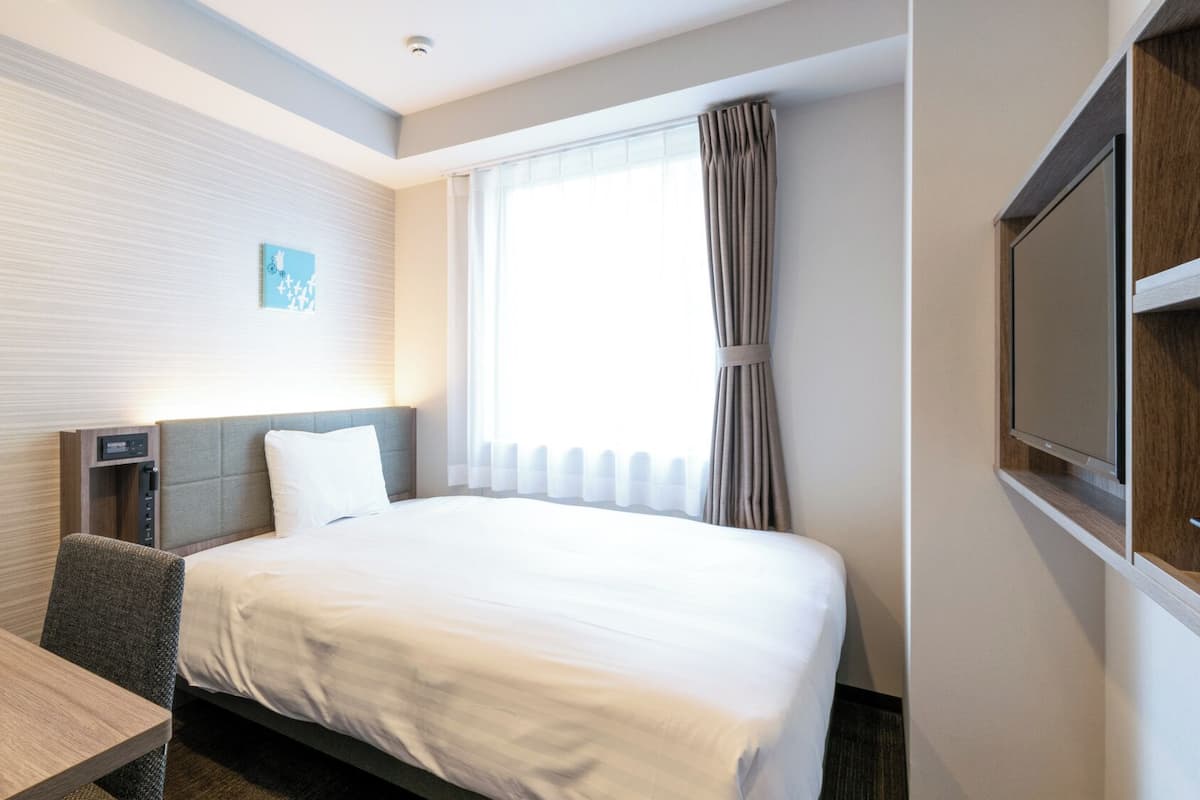 A hotel room with a bed, a desk, a chair, a TV, and a window with curtains.