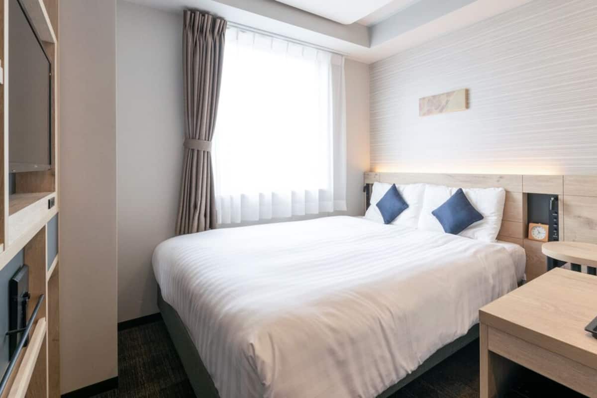 A hotel room with a bed, a bedside table with a tray, a wall-mounted clock, and a window with curtains.