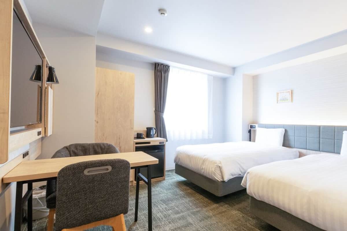 A hotel room with two beds, a desk, a chair, a wardrobe, a window with curtains, and a wall-mounted lamp.
