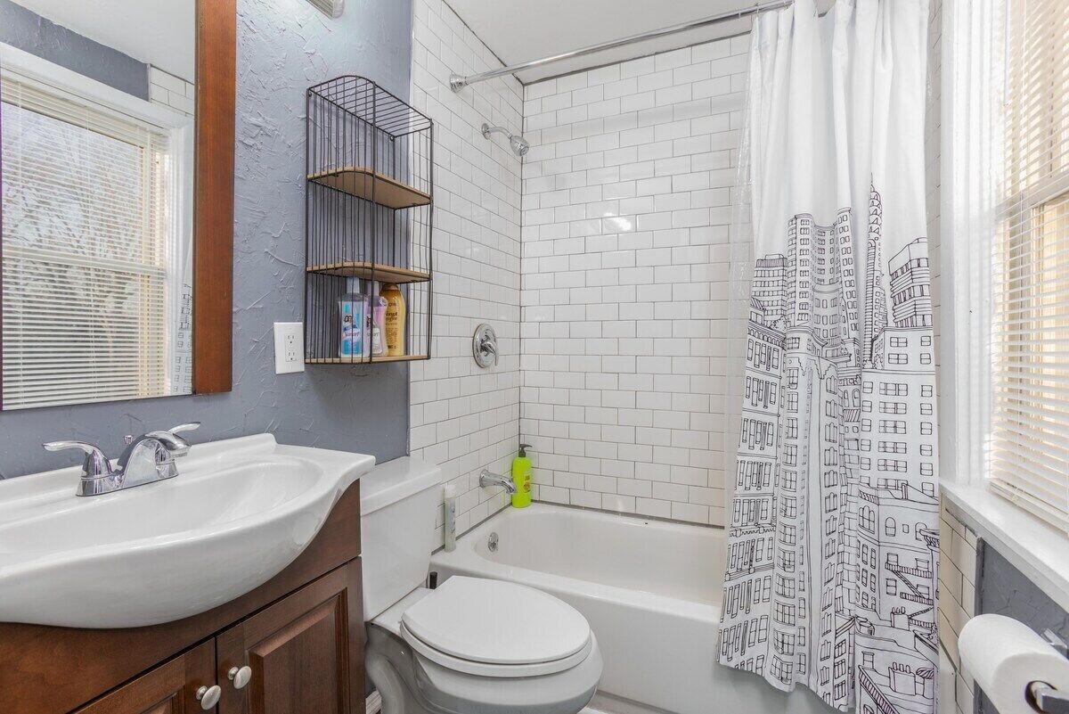 Een badkamer met een wit wastafelmeubel, toilet, bad en douchegordijn met een stadsgezicht.