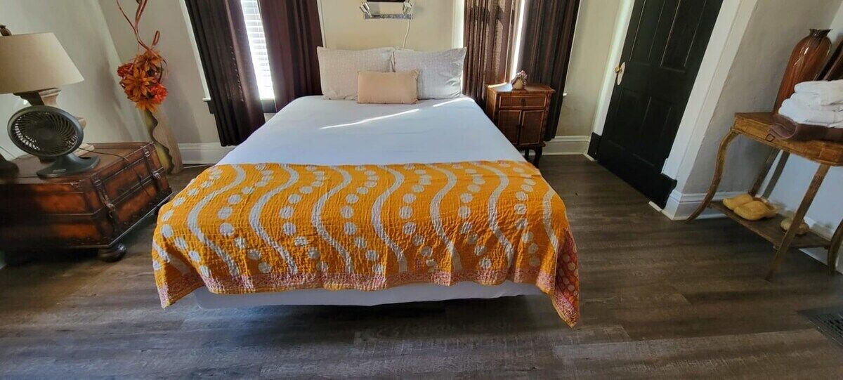 Ein ordentlich bezogenes Bett mit einer gemusterten Decke, ein Nachttisch mit einer Lampe und ein Holznachttisch mit einem Blumenstrauß.