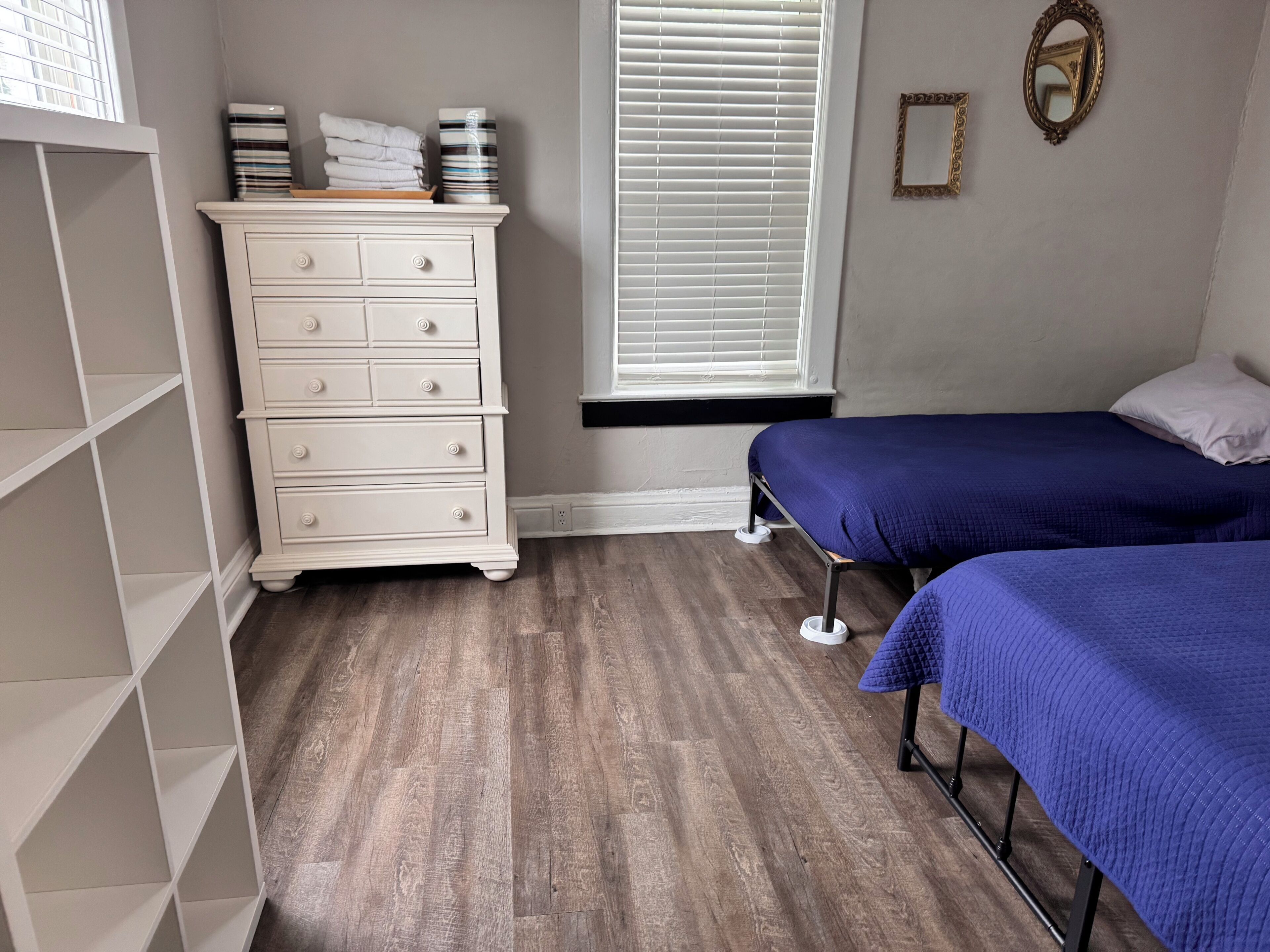 Une chambre avec une commode blanche, deux lits avec une literie bleue, une fenêtre avec des stores et un miroir sur le mur.