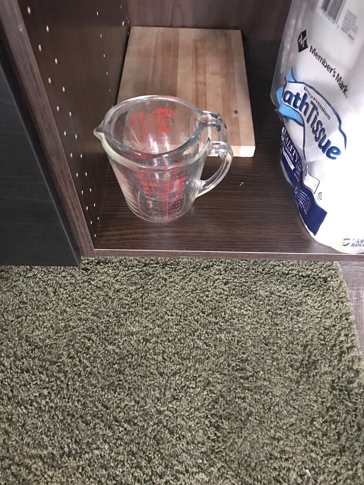 A measuring cup on a wooden surface, a package of toilet paper, and a green textured mat.