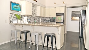 A modern kitchen with a granite countertop, white cabinets, and a stainless steel oven. There are three bar stools and a potted plant on the counter.