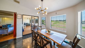 A spacious dining area with a wooden table and chairs, large windows with blinds, and a modern chandelier.