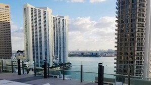 A rooftop terrace with white lounge chairs, a glass railing, and a view of a river and city skyline. - Fun Getaway in the Center of Downtown Miami , available for art Basel (Miami)