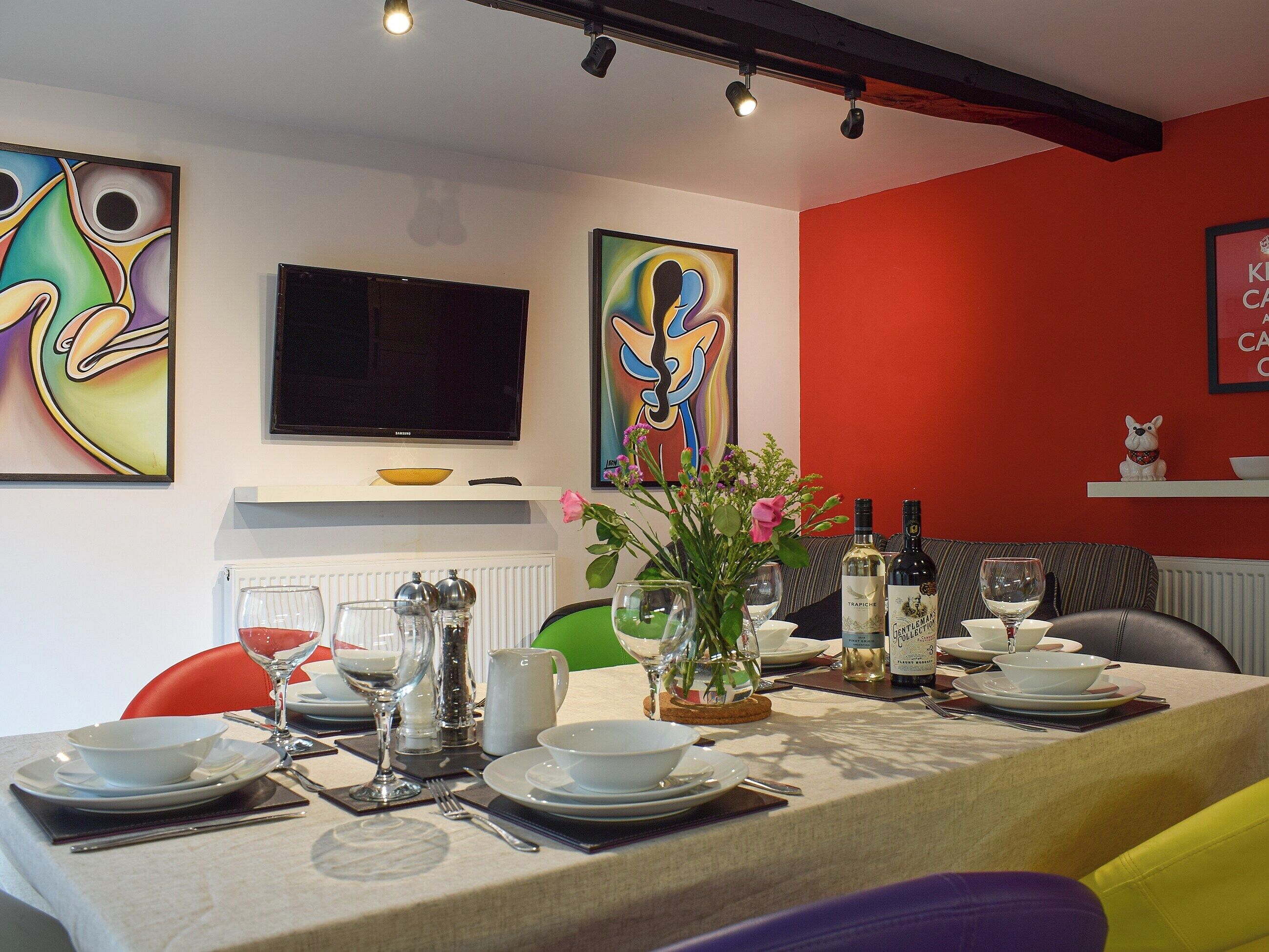 Un coin repas avec une table dressée pour quatre personnes, un téléviseur au-dessus et des œuvres d’art abstraites et colorées accrochées aux murs.