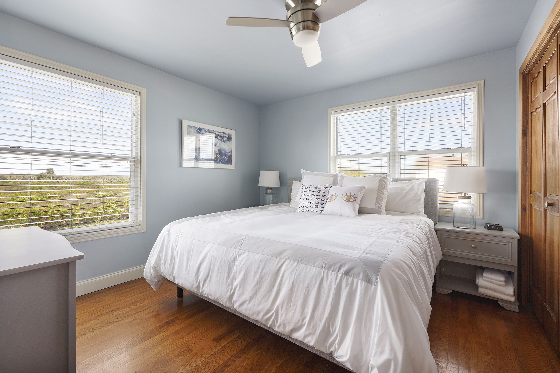 A bedroom with a bed, two bedside tables with lamps, a ceiling fan, and large windows.