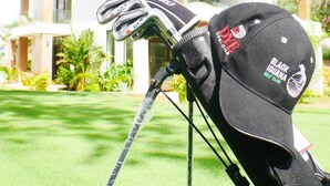 A golf club head with a cap on a stand, set against a modern house with a red roof and large windows. - West Bay Golf Club & Villas (Roatan)