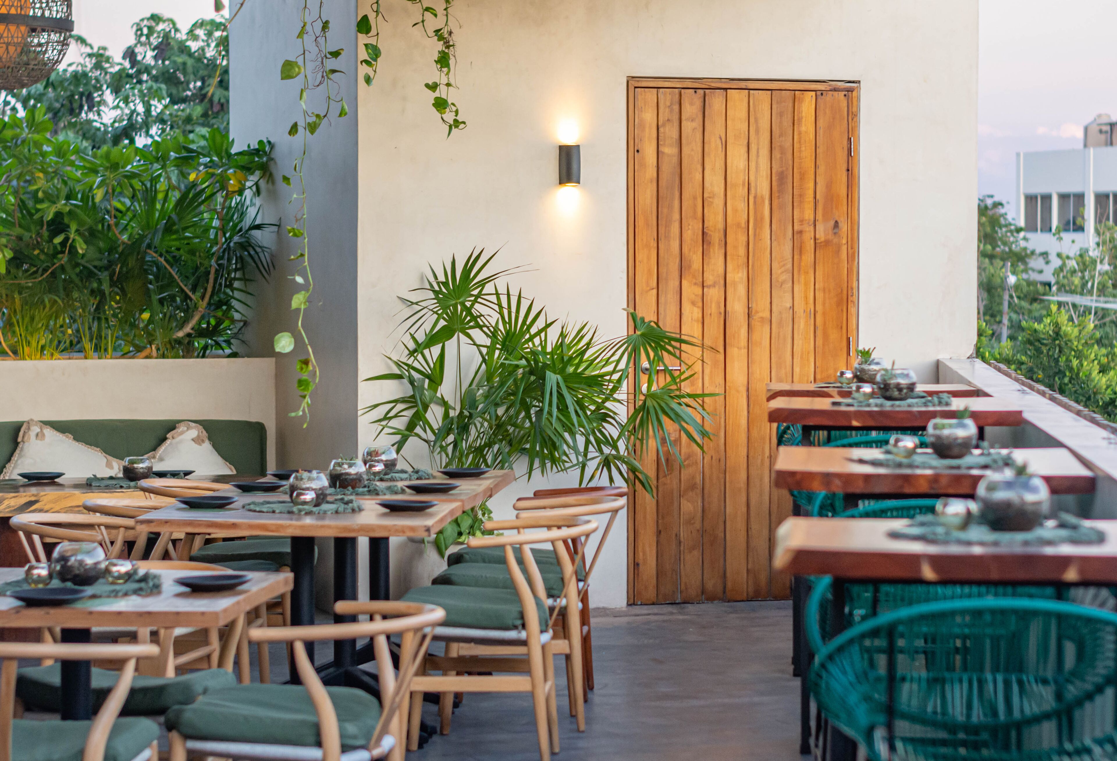 An outdoor dining area with wooden tables and chairs, surrounded by greenery and a wooden door.