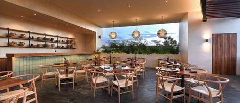 Un restaurant avec des tables et des chaises en bois, une étagère où sont exposées des poteries, et une vue sur la verdure par la fenêtre.