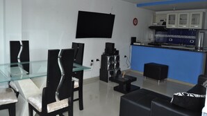 A modern kitchen with a blue accent wall, a dining area with glass-top table and black chairs, and a black sofa. - Las Margaritas (Cali)