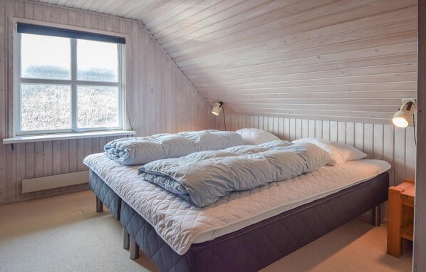 A bedroom with a sloped ceiling, a large window, two bedside lamps, and a bed with a quilted duvet. - Amazing home in Blåvand with sauna (Blåvand)