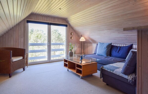 A cozy room with a sloped ceiling, a wicker sofa, a glass coffee table, and a large sliding glass door leading to an outdoor area. - Amazing home in Blåvand with sauna (Blåvand)