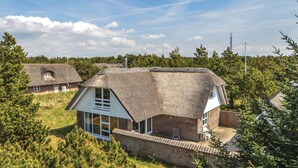 Une maison avec un toit de chaume et des murs en briques, entourée d’arbres et sous un ciel dégagé.